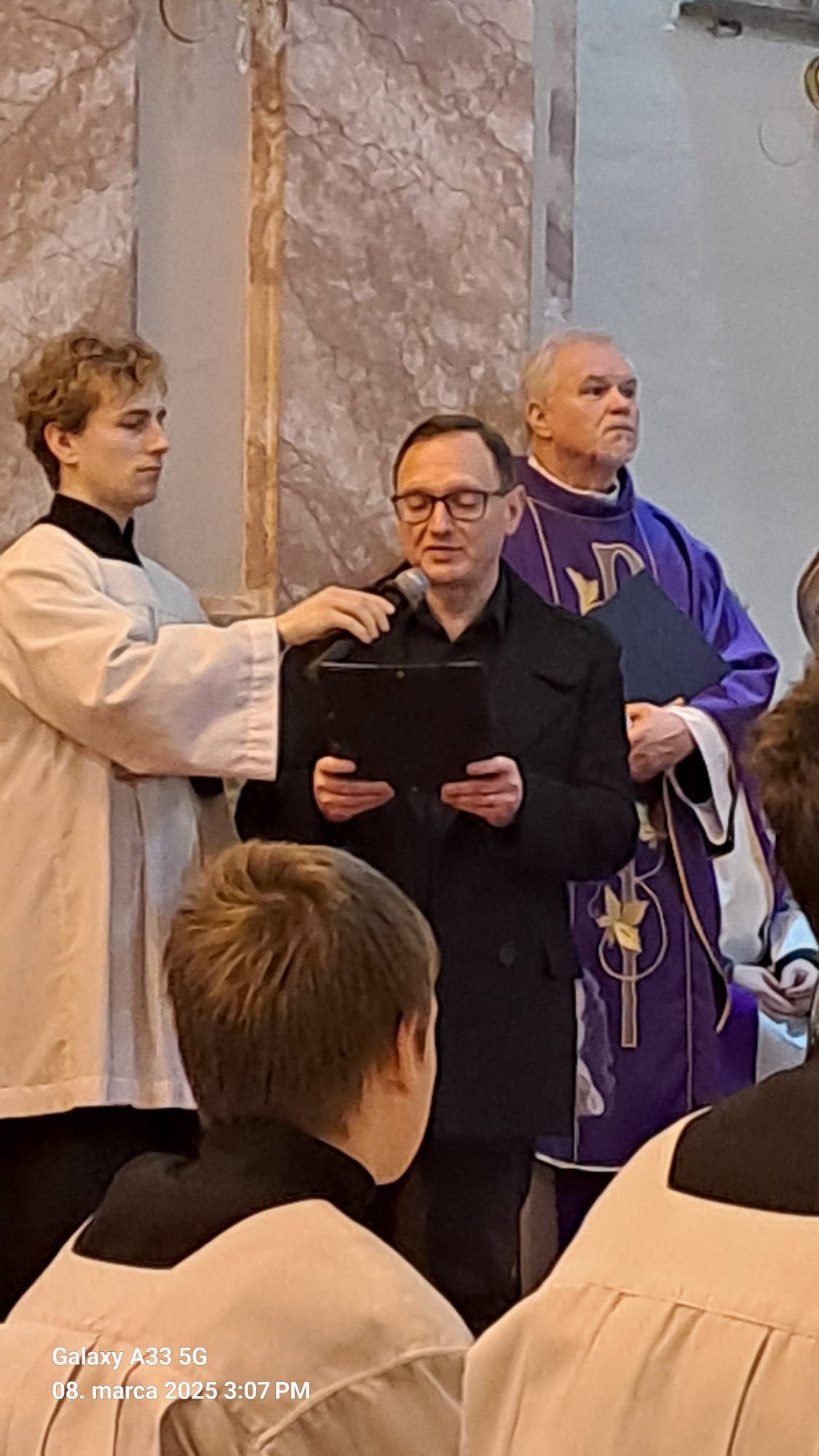 Pohľad do interiéru kostola počas pohrebnej omše Mons. Mariána Feduša v Rišňovciach, 8. marca 2025. Okolo oltára kňazi v liturgických rúchach fialovej farby a miništranti, na pozadí oltár s bohatou výzdobou a svietiacim lustrom nad ním.