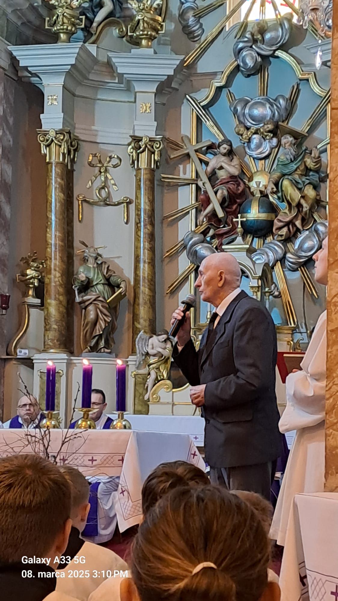 Pohľad do interiéru kostola počas pohrebnej omše Mons. Mariána Feduša v Rišňovciach, 8. marca 2025. Okolo oltára kňazi v liturgických rúchach fialovej farby a miništranti, na pozadí oltár s bohatou výzdobou a svietiacim lustrom nad ním.