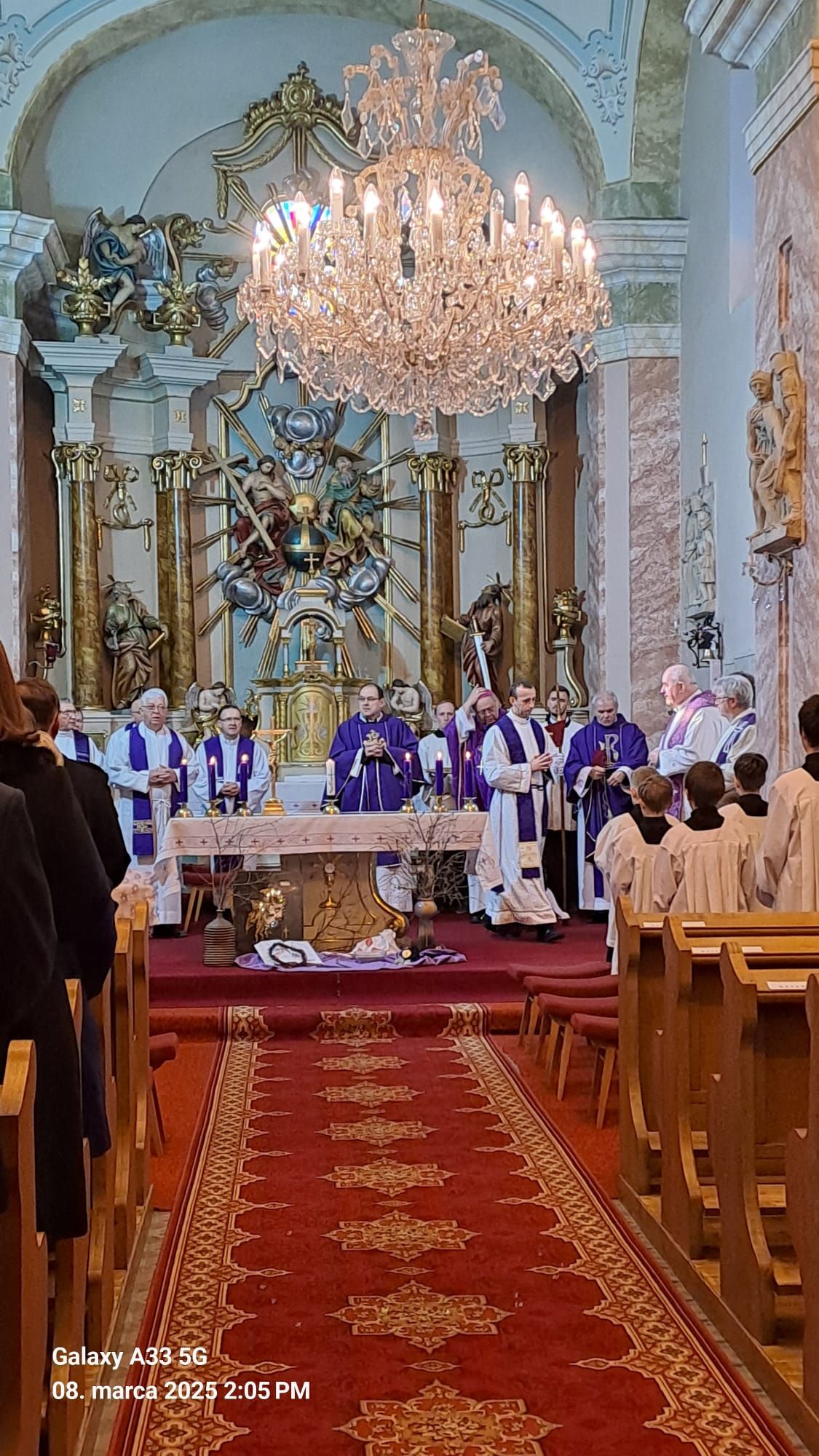 Pohľad do interiéru kostola počas pohrebnej omše Mons. Mariána Feduša v Rišňovciach, 8. marca 2025. Okolo oltára kňazi v liturgických rúchach fialovej farby a miništranti, na pozadí oltár s bohatou výzdobou a svietiacim lustrom nad ním.