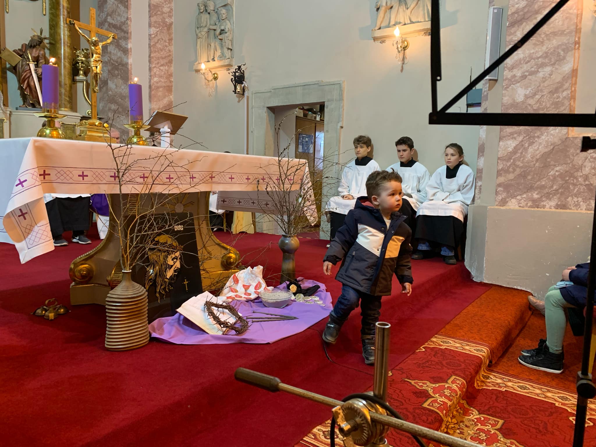 Malý chlapec stojí pri oltári v kostole v Rišňovciach, obklopený fialovými sviecami a liturgickými predmetmi, v pozadí miništranti v bielych rúchach
