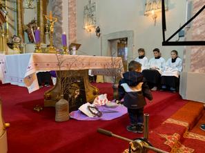 Malý chlapec stojí pri oltári v kostole v Rišňovciach, obklopený fialovými sviecami a liturgickými predmetmi, v pozadí miništranti v bielych rúchach