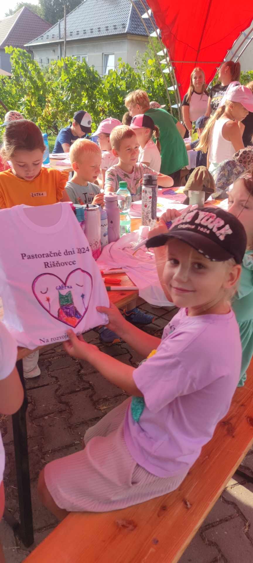 Rozložený stan so stolmi a lavičkami kde sedia deti s rodičmi a kňazom stojacími popri stanu.