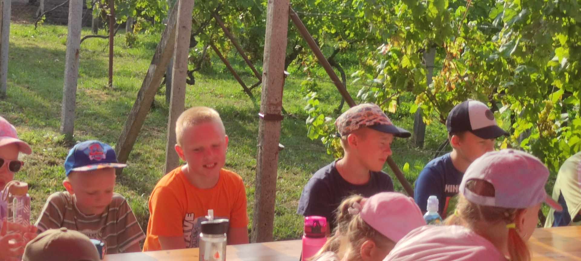 Rozložený stan so stolmi a lavičkami kde sedia deti s rodičmi a kňazom stojacími popri stanu.