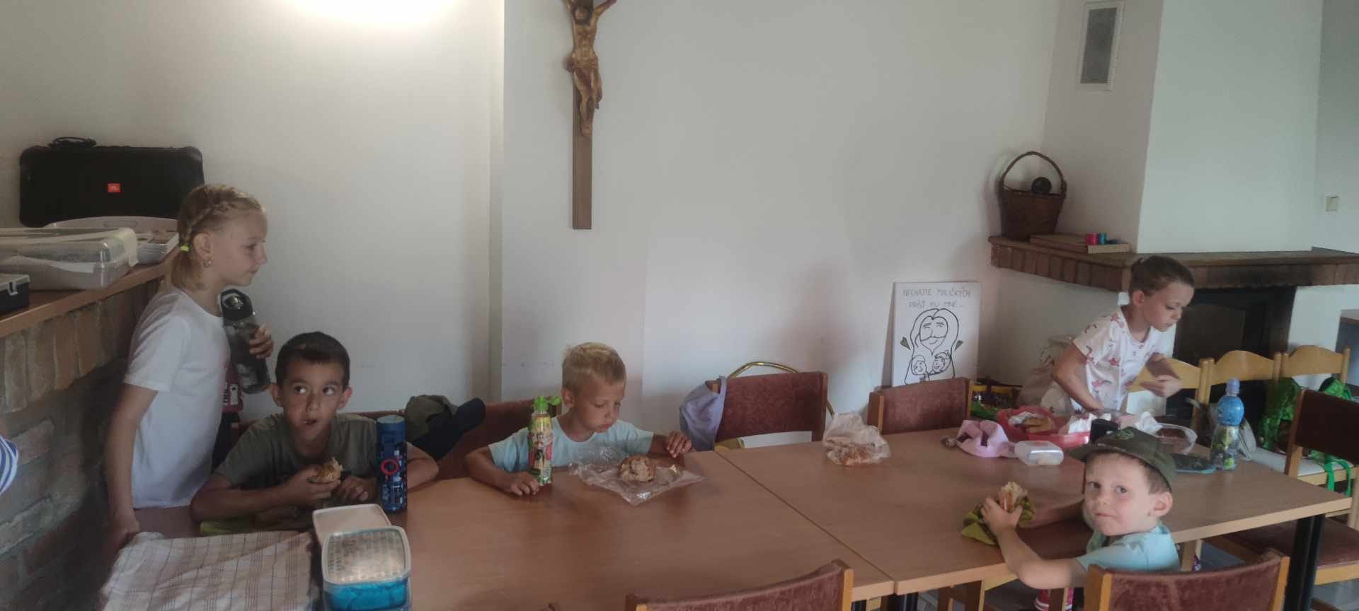 Rozložený stan so stolmi a lavičkami kde sedia deti s rodičmi a kňazom stojacími popri stanu.