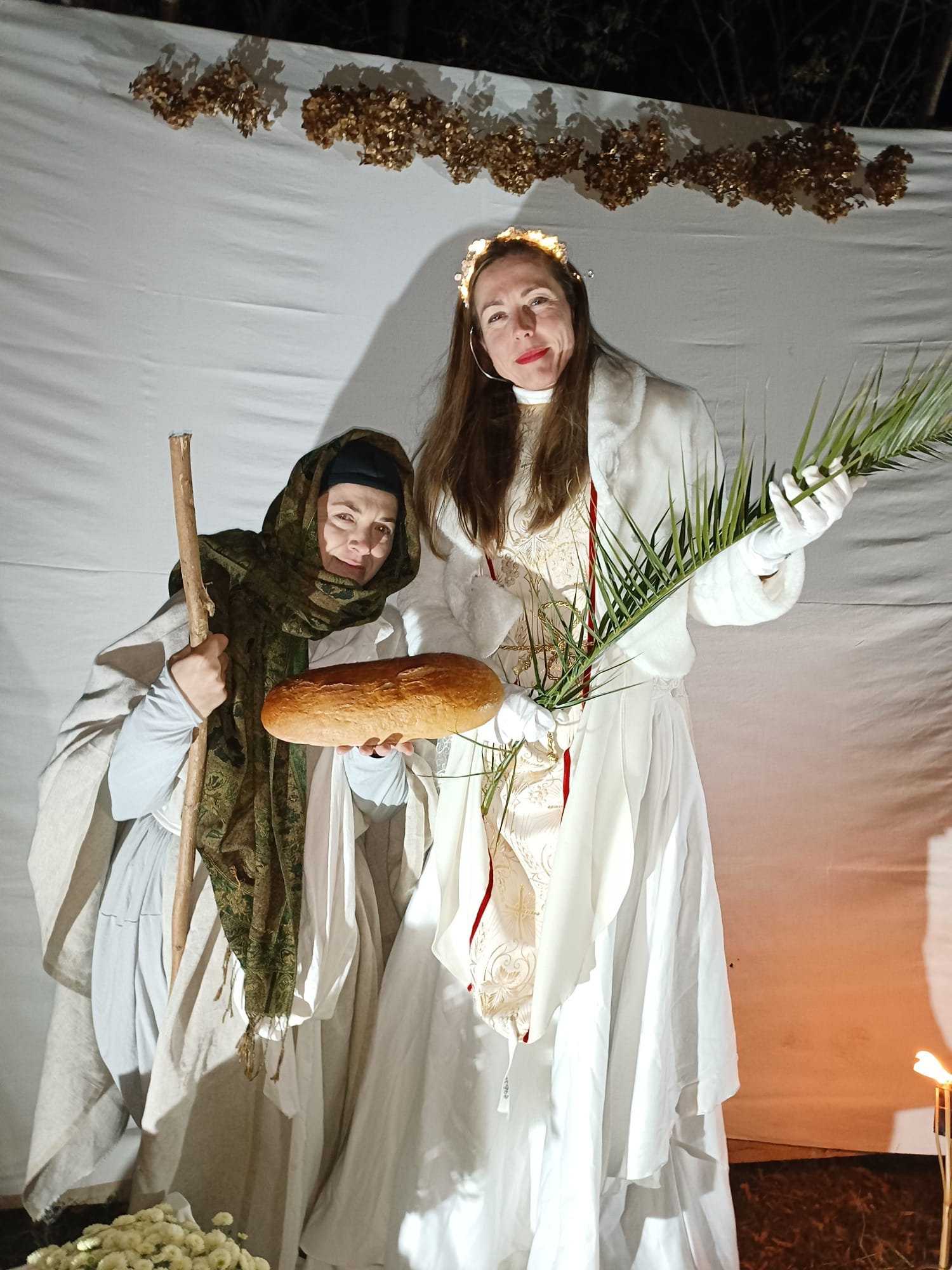 Skupinová fotografia účastníkov podujatia Svätí za dedinou v kostýmoch anjelov, svätcov a iných biblických postáv, nasvietená vonkajšími svetielkami, urobená v noci pod holým nebom.