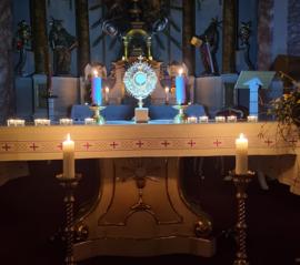 Adventná adorácia vo Farskom kostole Najsvätejšej Trojice v Rišňovciach, s vyloženou Eucharistiou na osvetlenom oltári, adventným vencom a zapálenými sviečkami v prítmí kostola